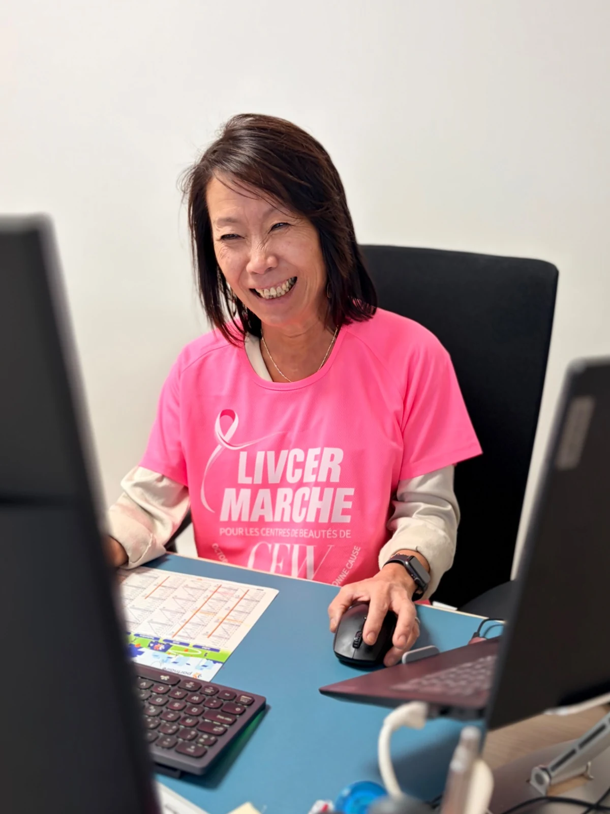 Une collaboratrice en t-shirt rose, souriante à son poste de travail.